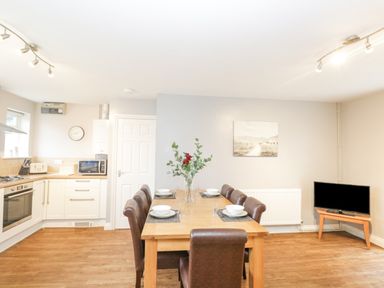 A dining area with a wooden table and six brown chairs set with plates and glasses next to a kitchen and a television on a stand at The Brambles in Monmouth