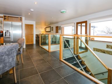 A kitchen and dining area with gray tiled floor a glass staircase railing wooden doors and stainless steel refrigerator at Swyn Llyr in Rhosneigr