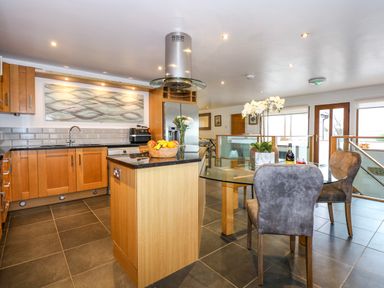 A kitchen with wooden cabinets an island with fruit a glass dining table with chairs and a glass door at Swyn Llyr in Rhosneigr