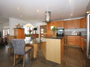 A kitchen with wooden cabinets a glass dining table and a kitchen island with flowers at Swyn Llyr in Rhosneigr