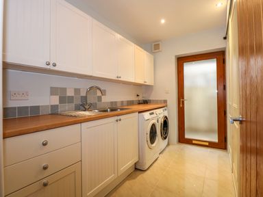 A utility room with white cabinets a wooden countertop a sink washing machine and dryer and a wooden door with frosted glass at Swyn Llyr in Rhosneigr