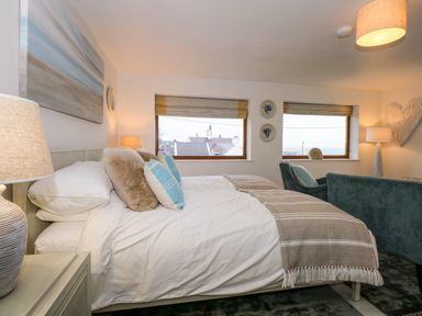 A bedroom with a made bed with pillows and throw blankets two windows two lamps a chair and a sofa at Swyn Llyr in Rhosneigr