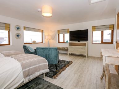A bedroom with a bed a blue armchair a bench a television on a cabinet and blinds on windows at Swyn Llyr in Rhosneigr