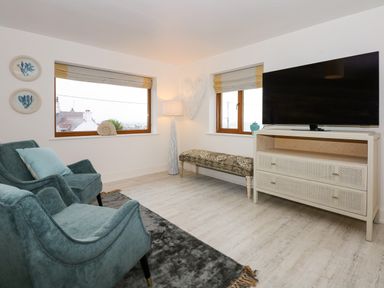 A living room with two upholstered chairs a flat screen tv on a cabinet and windows showing rooftops at Swyn Llyr in Rhosneigr