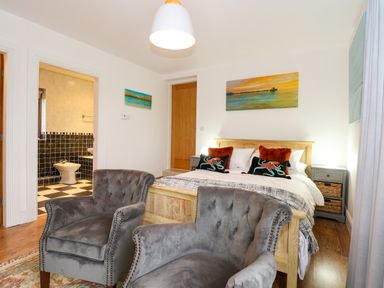 A bedroom with a bed pillows two gray armchairs paintings and a door leading to a bathroom at Swyn Llyr in Rhosneigr