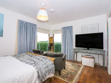 A bedroom with a bed a gray chair a large TV on a console table and blue curtains at Swyn Llyr in Rhosneigr