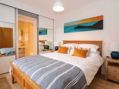 A bedroom with a wooden bed frame white and striped bedding orange pillows bedside tables with lamps mirrored closet doors and a painting above the bed at Swyn Llyr in Rhosneigr