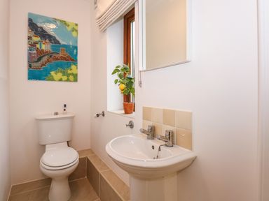 A bathroom with a toilet and a sink next to a window with a potted plant and a colorful wall painting at Swyn Llyr in Rhosneigr