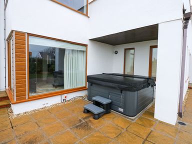 An outdoor patio with a covered hot tub and steps next to large windows at Swyn Llyr in Rhosneigr