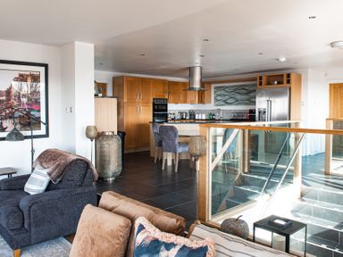 An open plan living area with a kitchen featuring wooden cabinets and a dining area near a staircase with glass railing at Swyn Llyr in Rhosneigr