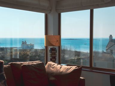 A living room corner with a brown sofa and a lamp by large windows overlooking the sea and nearby buildings at Swyn Llyr in Rhosneigr