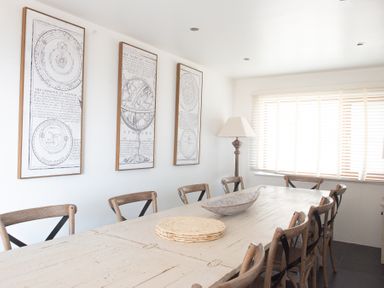 A dining room with a large wooden table surrounded by chairs a floor lamp three framed maps on the wall and window blinds at Swyn Llyr in Rhosneigr