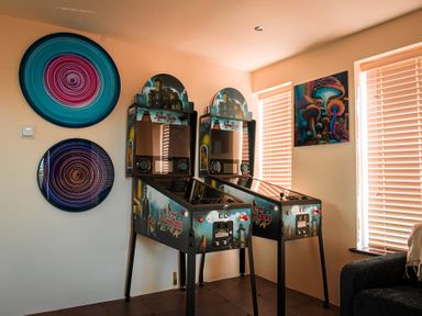 A room with two Addams Family pinball machines two circular wall art pieces and a painting of mushrooms near windows with blinds at Swyn Llyr in Rhosneigr