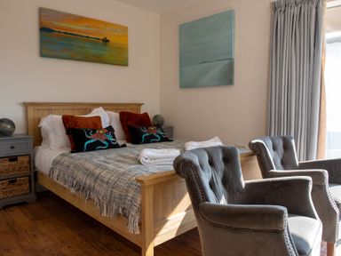 A bedroom with a wooden bed, two bedside tables, two gray armchairs, and paintings on the walls at Swyn Llyr in Rhosneigr