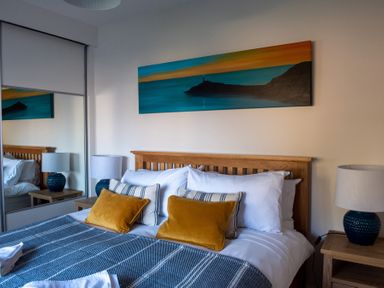A bedroom with a wooden bed frame blue and white bedding yellow pillows framed artwork above the bed and bedside tables with lamps at Swyn Llyr in Rhosneigr