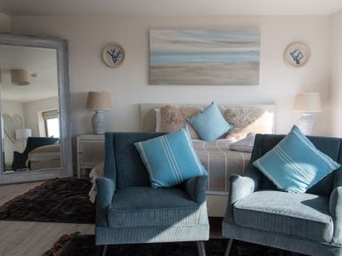 A bedroom with two blue armchairs and pillows a bed with cushions and lamps on side tables at Swyn Llyr in Rhosneigr