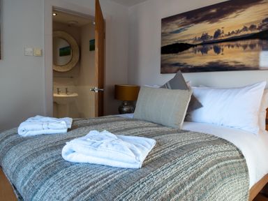 A bedroom with a bed with gray and beige cushions and towels on a gray blanket and an open door to a bathroom at Swyn Llyr in Rhosneigr