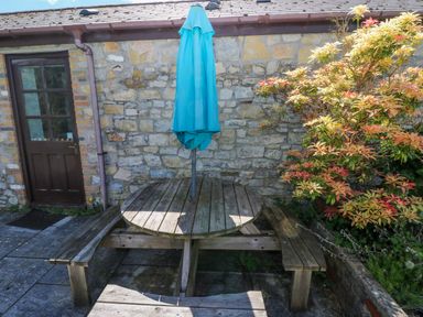 An outdoor area with a round wooden picnic table and benches with a closed blue umbrella next to a stone wall and a door with a brown frame at Bluebell Cottage in St Brides-super-Ely near Cardiff