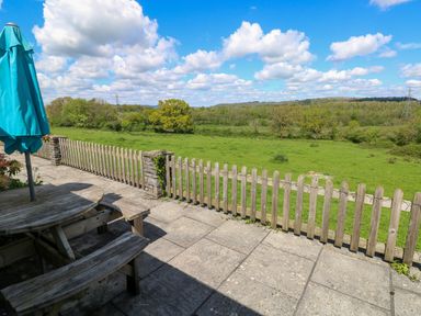 A patio with a wooden table bench set and umbrella overlooking a fenced green field at Bluebell Cottage in St Brides-super-Ely near Cardiff