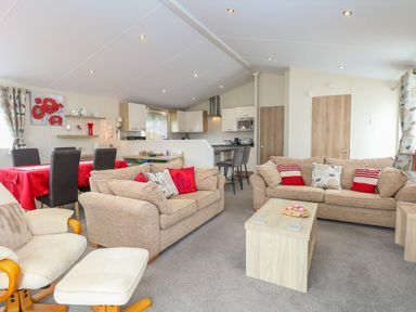 A living room with beige sofas red and patterned cushions a wooden coffee table and dining table with red tablecloth at 21 Norman Drive Lodge in Burgh Castle