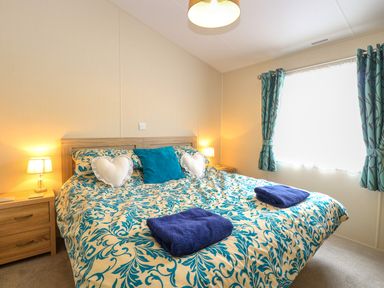 A bedroom with a double bed two bedside tables lamps folded towels and a window with curtains at 21 Norman Drive Lodge in Burgh Castle