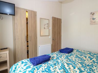A bedroom with a bed covered in a patterned blanket and folded towels on top a wardrobe a wall-mounted television and a wooden door at 21 Norman Drive Lodge in Burgh Castle