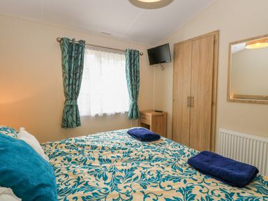 A bedroom with a bed covered in blue and beige patterned bedding two folded blue towels on the bed a window with blue curtains a wooden wardrobe a small wooden nightstand a wall-mounted television at 21 Norman Drive Lodge Burgh Castle