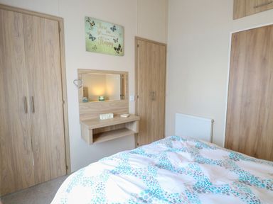 A bedroom with a bed with a patterned duvet a wall-mounted vanity mirror wooden wardrobes and a wall sign at 21 Norman Drive Lodge in Burgh Castle