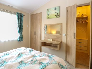 A bedroom with a bed a window with curtains a wall mirror and an open door to a closet at 21 Norman Drive Lodge Burgh Castle