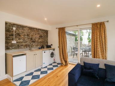 A kitchen area with washing machine fridge and sink next to sliding glass doors with yellow floral curtains and a blue sofa with pillows at The Farm Cottage in Athy County Kildare