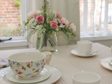 A dining table set with floral patterned bowls white cups and a vase with flowers near a window at Ty Hafan in Mostyn
