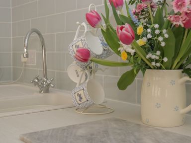 A kitchen countertop with a sink faucet a cup holder with cups and a flower vase with pink and yellow flowers at Ty Hafan in Mostyn