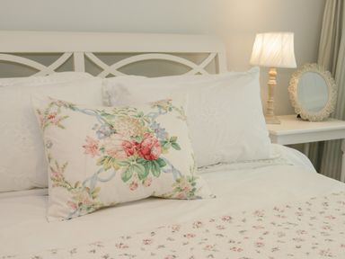 A bed with white pillows and a floral pillow in front next to a nightstand with a lamp and a round mirror at Ty Hafan in Mostyn