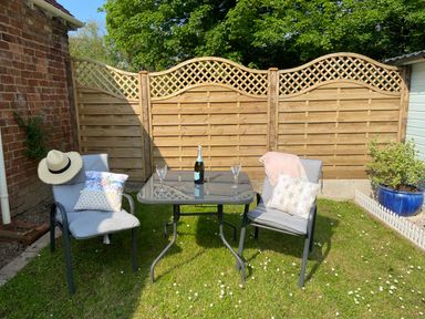 An outdoor patio area with two cushioned chairs, a glass table with a bottle and two glasses, a wooden fence, a potted plant, and a brick wall at Ty Hafan in Mostyn