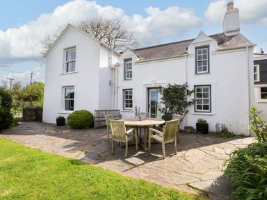 A white house with windows and a chimney with a stone patio with a round table and chairs and green lawn at Bryn Tirion in Morfa Nefyn