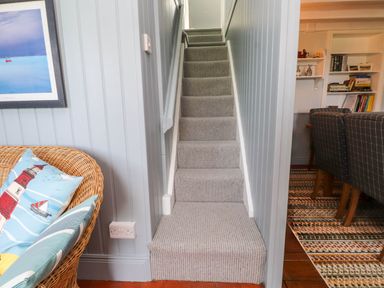 A narrow carpeted staircase between gray paneled walls with a wicker chair and cushions to the left and a dining area with chairs and bookshelves to the right at Bryn Tirion in Morfa Nefyn