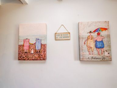 Two framed beach-themed artworks and a hanging sign on a wall at Bryn Tirion Morfa Nefyn