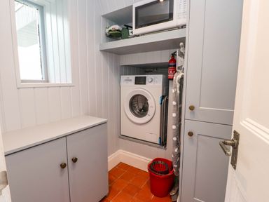 A laundry room with a washing machine microwave cabinet an ironing board a bucket and mop at Bryn Tirion in Morfa Nefyn