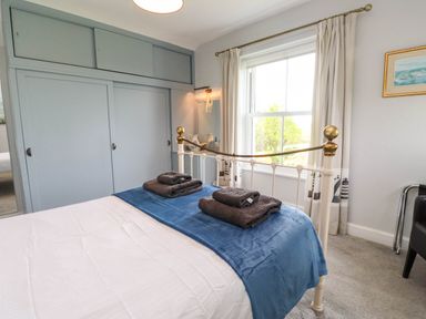 A bedroom with a white metal bed frame blue throw blanket and folded towels on the bed built in wardrobes a window with curtains and a carpeted floor at Bryn Tirion in Morfa Nefyn