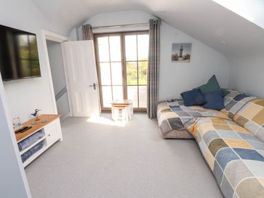 A room with a wall-mounted television a white wooden cabinet a sofa with a checkered cover and pillows a set of small tables and glass doors covered by curtains at Bryn Tirion in Morfa Nefyn