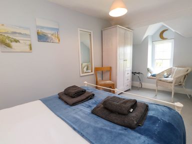 A bedroom with a bed, folded towels, a wardrobe, chair, mirror, wicker chair, and a window seat at Bryn Tirion in Morfa Nefyn