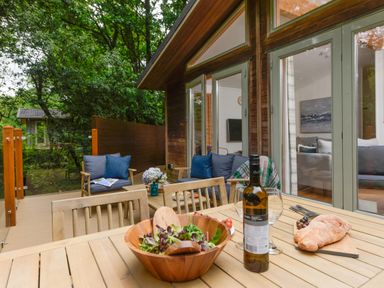 An outdoor wooden patio with wooden chairs a table with salad bread wine glasses and sofas with cushions at 10 Stonerush Valley Lanreath near Looe
