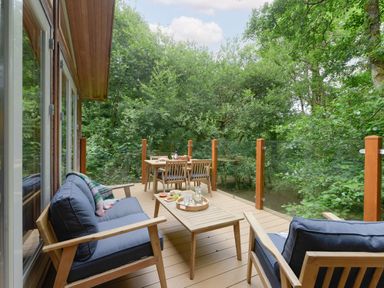 An outdoor wooden deck with cushioned chairs a table with drinks and food surrounded by trees at 10 Stonerush Valley Lanreath near Looe