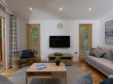 A living room with a television on the wall two windows with blinds a sofa a chair two tables and a rug at 10 Stonerush Valley Lanreath near Looe