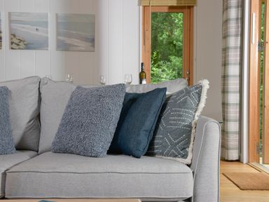 A grey sofa with three blue cushions in a living room with wooden framed glass doors and paintings on the wall at 10 Stonerush Valley in Lanreath near Looe