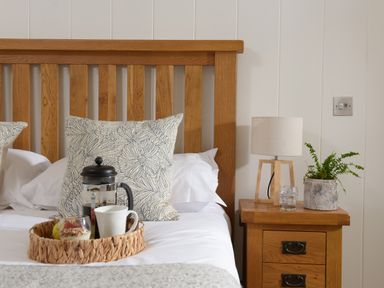 A wooden bed with patterned pillows and a tray with a coffee pot and cups next to a wooden nightstand with a lamp and plant at 10 Stonerush Valley Lanreath near Looe