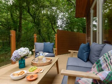 An outdoor seating area with a wooden table holding snacks and drinks surrounded by cushions on a deck at 10 Stonerush Valley in Lanreath near Looe