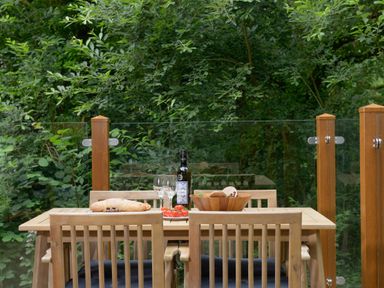 An outdoor wooden dining table with chairs a bottle of wine two wine glasses bread and a bowl on the table with green trees in the background at 10 Stonerush Valley Lanreath near Looe