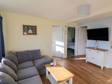 A living room with a gray sofa a wooden coffee table a wall-mounted television and an open door showing a bedroom at The Sail Loft in Norton Park near Dartmouth
