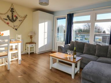 A living room with a gray sofa a wooden coffee table with wine bottle and glasses a dining table with chairs a wardrobe and nautical decor at The Sail Loft in Norton Park near Dartmouth